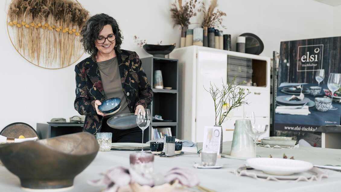 Eine Frau dekoriert einen Tisch mit Geschirr und Gläsern. Im Hintergrund sind dekorative Elemente und ein Werbebanner sichtbar. | © vulkanland.at/bergmann
