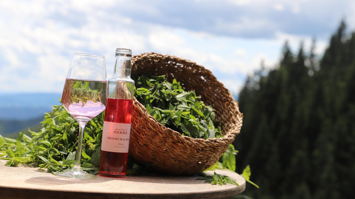 Ein Glas mit rosa Basilikum-Minz-Getränk und eine Flasche Sirup stehen neben einem Weidenkorb voller frischer Kräuter, vor einer malerischen Berglandschaft im Kräutergarten gratia HERBA in Geistthal-Södingberg. | © Anna Höller