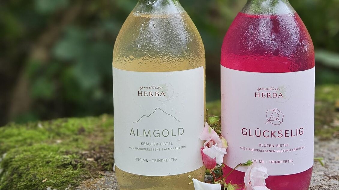 Zwei Flaschen Kräutersäfte von Gratia HERBA – Almgold und Glückselig – stehen auf einem moosbedeckten Stein, umgeben von frischen Blumen in Geistthal-Södingberg.  | © Anna Höller