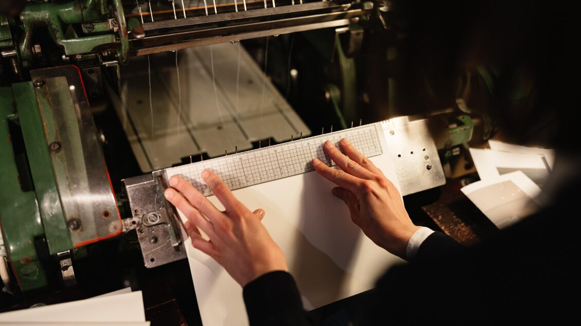 Hände richten einen Papierbogen an einer historischen Maschine in der Buchbinderei maarble in Graz aus. | © Bernd Grosseck
