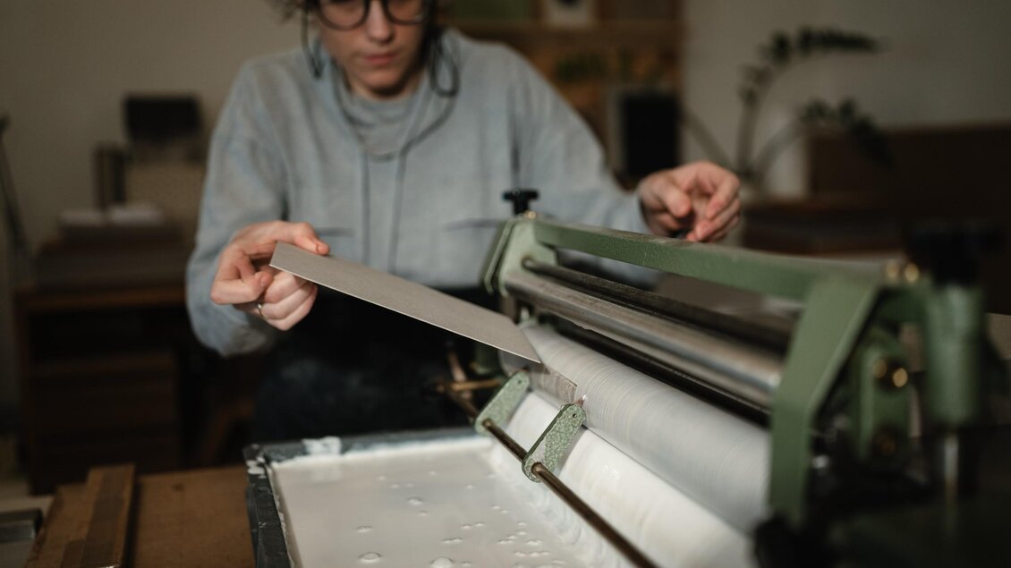 Eine Buchbinderin führt einen Karton durch das Leimbad einer Maschine in der Buchbinderei maarble in Graz. | © Bernd Grosseck