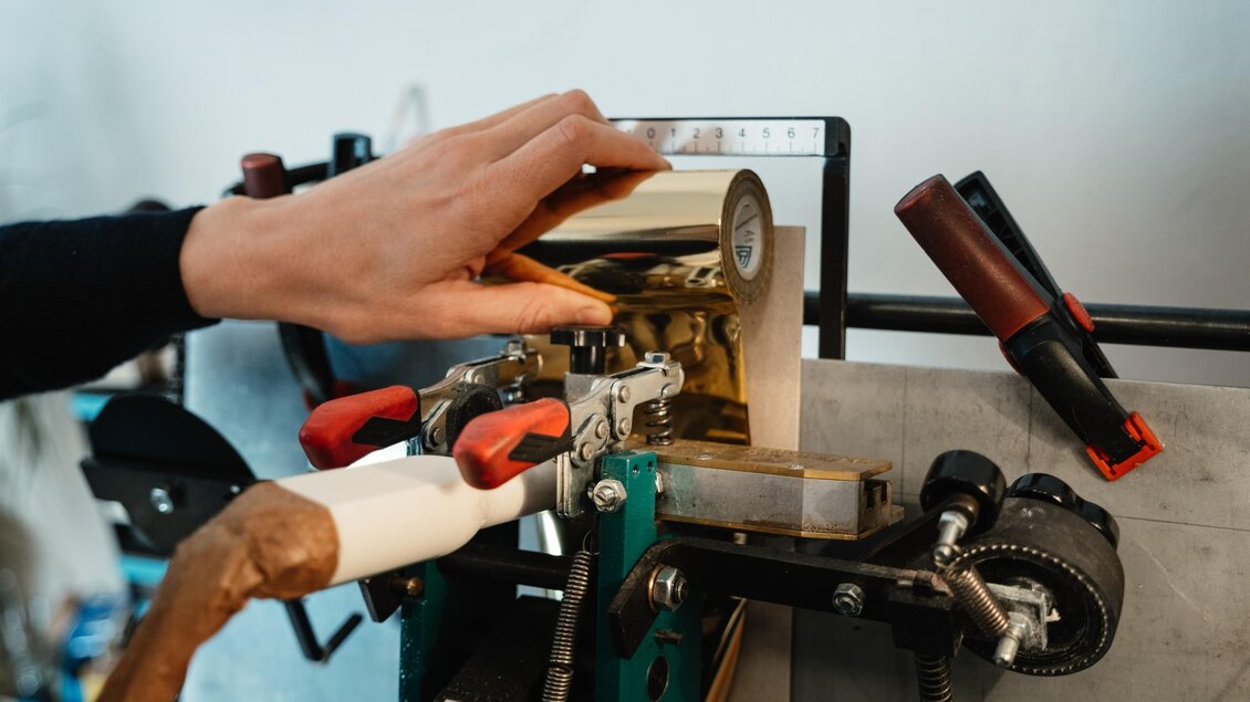 Eine Hand führt eine Rolle mit Blattgold an einer Maschine in der Buchbinderei maarble in Graz. | © Bernd Grosseck