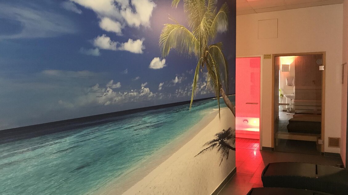 Ein entspannender Raum mit einem Strandmotiv an der Wand. Im Hintergrund sind Wellness-Einrichtungen sichtbar. | © Schreiber Daniel