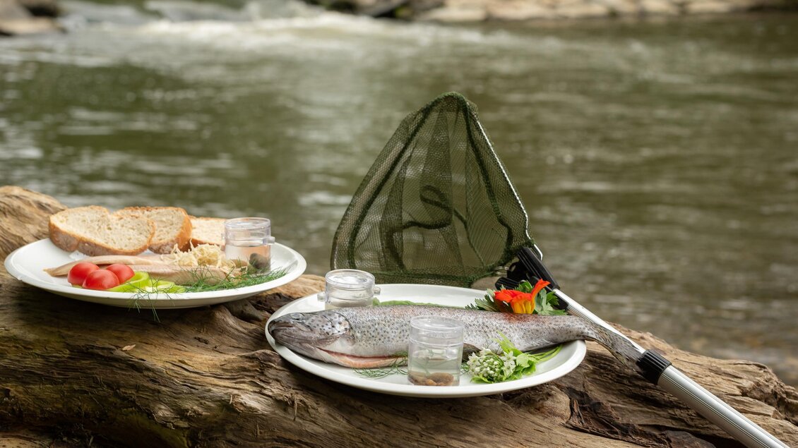 Ein Holztisch am Fluss mit zwei Tellern. Auf einem Teller liegt ein frischer Fisch mit Beilagen, während der andere mit Brot und Gemüse dekoriert ist. | © RM SW GmbH | webquartier.at