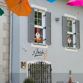 Ein charmantes Gebäude mit grauer Fassade und bunten Regenschirmen, die über dem Eingang hängen. Vor dem Gebäude stehen elegante Stehtische und Pflanzen in Töpfen. | © TVB Südsteiermark