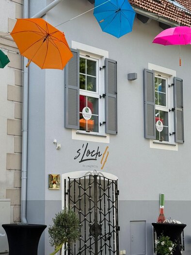 Ein charmantes Gebäude mit grauer Fassade und bunten Regenschirmen, die über dem Eingang hängen. Vor dem Gebäude stehen elegante Stehtische und Pflanzen in Töpfen. | © TVB Südsteiermark