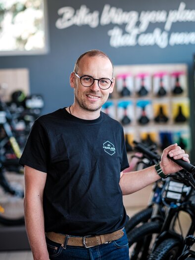 A smiling man is standing in a bicycle accessories store. In the background, various bicycles and accessories are visible. | © s´Radl Eck Gollinger