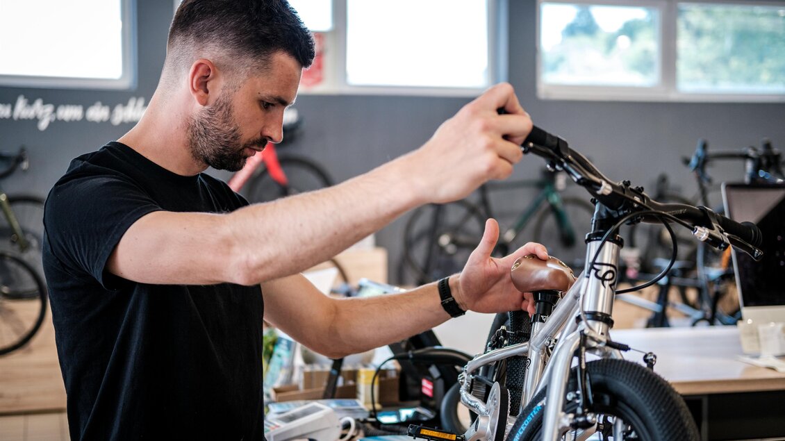 Ein Mann arbeitet an einem Fahrrad in einer Werkstatt. Im Hintergrund sind weitere Fahrräder und Werkzeuge sichtbar. | © s´Radl Eck Gollinger