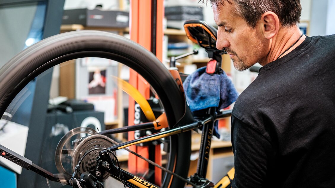 Ein Mann prüft das Hinterrad eines Fahrrads in einer Werkstatt. Das Fahrrad ist orange und schwarz, mit Werkstattutensilien im Hintergrund. | © s´Radl Eck Gollinger