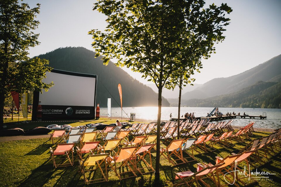 Eine Open-Air-Kinoaufführung am See mit bunten Liegestühlen. Im Hintergrund sind Berge und ein klarer Himmel zu sehen. | © Fred Lindmoser