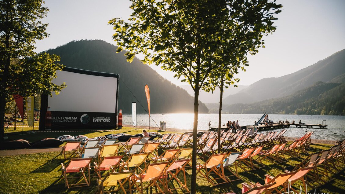 Eine Open-Air-Kinoaufführung am See mit bunten Liegestühlen. Im Hintergrund sind Berge und ein klarer Himmel zu sehen. | © Fred Lindmoser