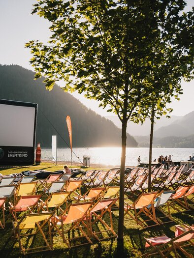 Eine Open-Air-Kinoaufführung am See mit bunten Liegestühlen. Im Hintergrund sind Berge und ein klarer Himmel zu sehen. | © Fred Lindmoser