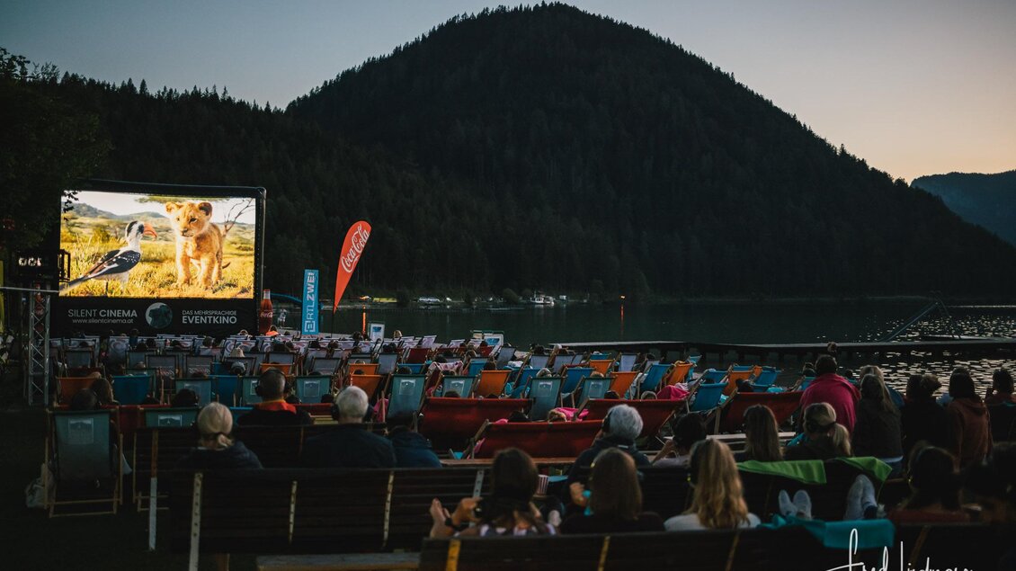 Eine Freiluftkinoveranstaltung am Abend mit einem großen Bildschirm. Im Hintergrund sind Berge und der See sichtbar, während die Zuschauer auf Stühlen sitzen und den Film genießen. | © Fred Lindmoser