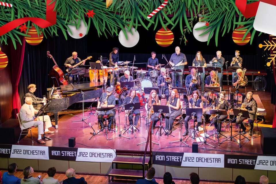 Ein großes Jazz-Orchester spielt auf einer Bühne mit verschiedenen Instrumenten. Das Publikum sitzt im Hintergrund und genießt die Darbietung. | © ISJO