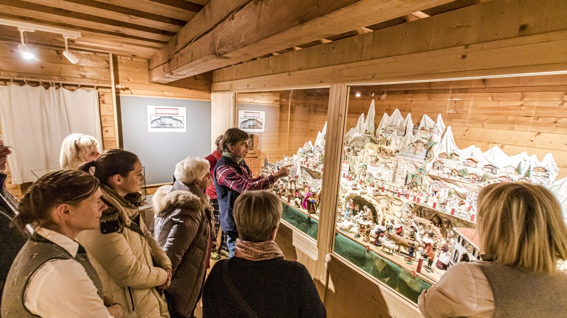 Eine Gruppe von Menschen betrachtet eine detailreiche Weihnachtskrippe in einem Ausstellungsraum. Sie stehen vor der Glasvitrine und scheinen interessiert zuzuhören. | © Gerhard Pilz
