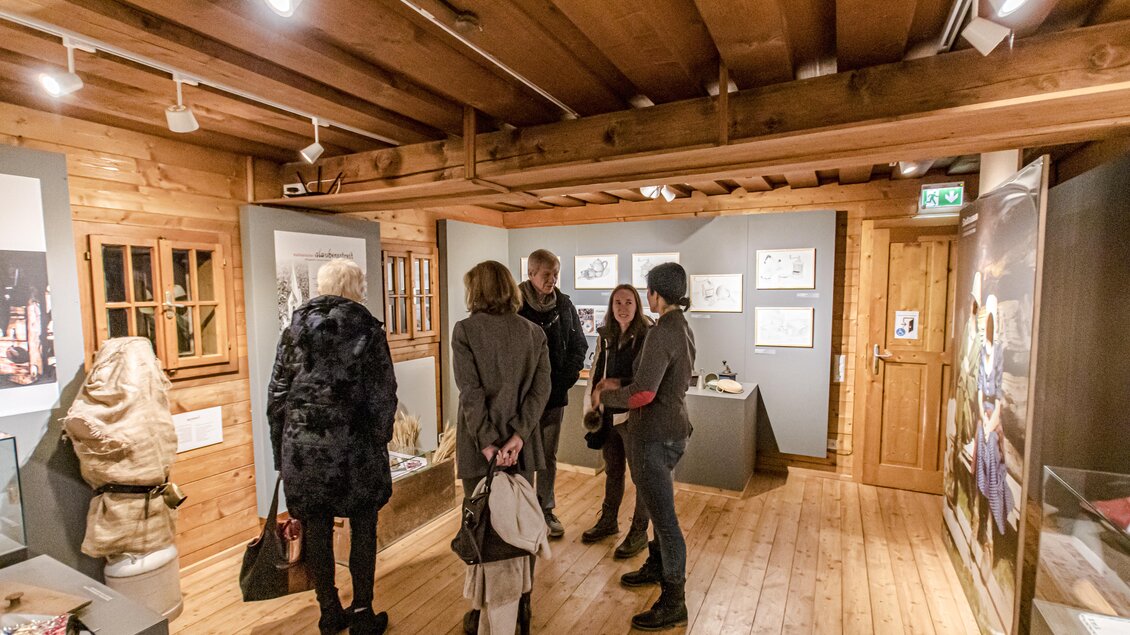 Eine Gruppe von Menschen steht in einem holzgetäfelten Raum und betrachtet Ausstellungsstücke. An den Wänden sind Bilder und Informationen zu sehen. | © Gerhard Pilz