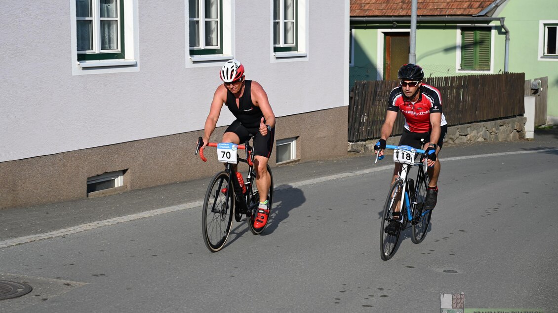 Zwei Radfahrer fahren auf einer ruhigen Straße vorbei an Häusern. Der erste Radfahrer trägt ein schwarzes Trikot, der zweite ein rotes Trikot. | © Marktgmeinde Kraubath