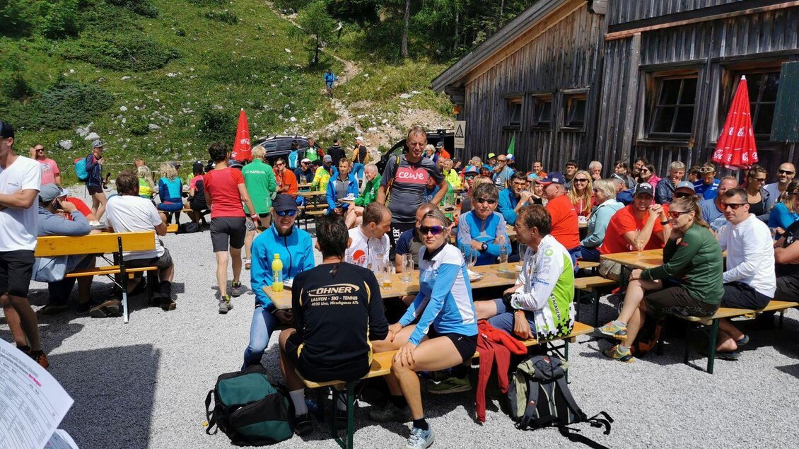 Eine lebhafte Veranstaltung im Freien mit vielen Menschen, die an Tischen sitzen. Im Hintergrund sind grüne Hügel und ein Holzgebäude zu sehen. | © Loitzl_Neuhuber