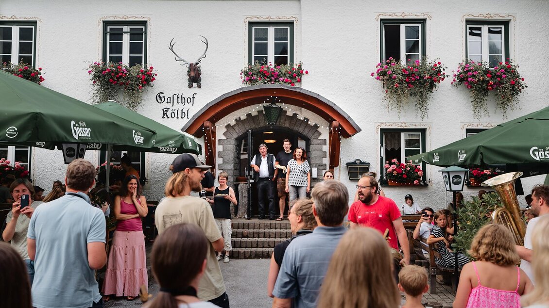 Eine belebte Gaststätte mit Menschen, die sich vor einem traditionellen Gebäude versammeln. Blumen schmücken die Fenster, während Gäste unter grünen Schirmen sitzen.