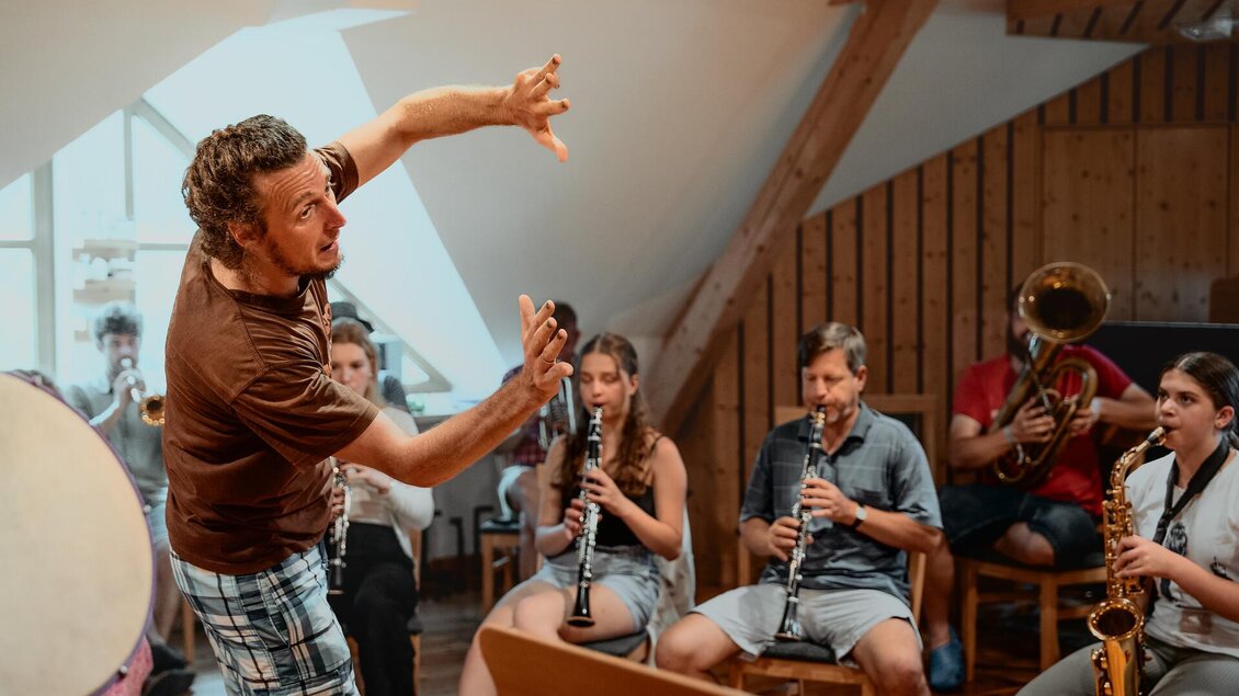 Eine Gruppe von Musikern probt in einem gemütlichen Raum. Ein Mann leitet mit viel Enthusiasmus die Proben.