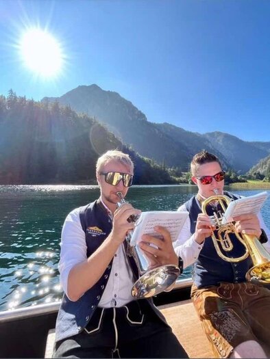 Two musicians are sitting in a boat on the water playing wind instruments. In the background, there are mountains and a clear sky. | © Andreas Swogoda