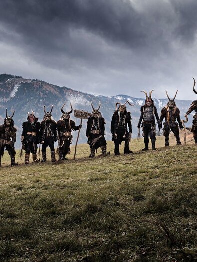 Eine Gruppe von mystisch gekleideten Figuren steht auf einer Wiese vor Bergen. Das Wetter ist bewölkt und die Szenerie hat eine dramatische Atmosphäre. | © Ehrenfels Pass Kammern