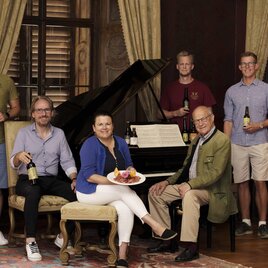 A group of five people stands around a piano holding bottles in their hands. A woman sits on a chair and presents a snack on a plate. | © Markenagentur Conterfei