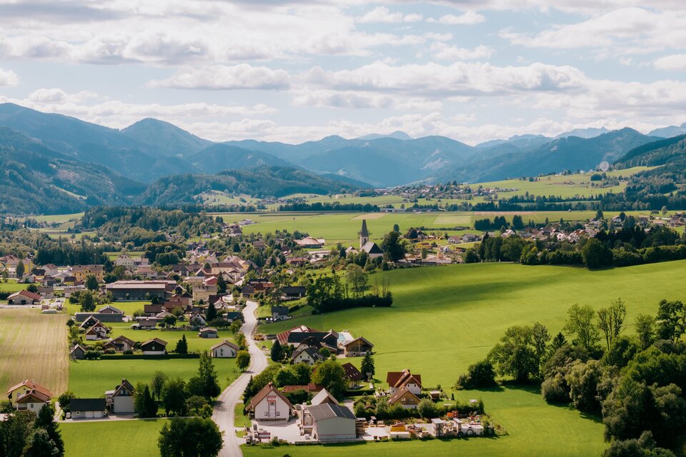 Eine malerische Landschaft mit grünen Feldern und kleinen Dörfern. Im Hintergrund erheben sich sanfte Berge unter einem bewölkten Himmel. | © Roman Königshofer
