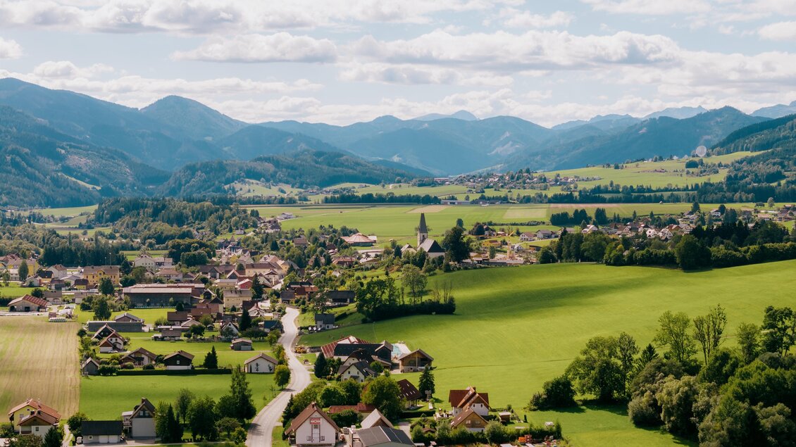 Eine malerische Landschaft mit grünen Feldern und kleinen Dörfern. Im Hintergrund erheben sich sanfte Berge unter einem bewölkten Himmel. | © Roman Königshofer