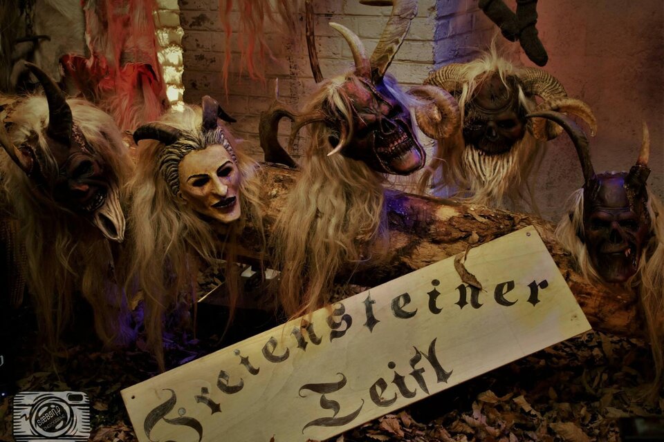 Eine Gruppe von bemalten Masken mit langen Haaren ist auf einem Tisch ausgestellt. Im Vordergrund steht ein Schild mit der Aufschrift „Zietensteiner Teifl“. | © Freiensteiner Teifl