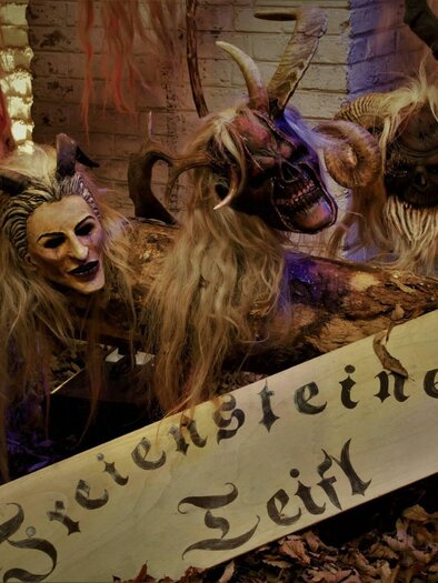 A group of painted masks with long hair is displayed on a table. In the foreground, there is a sign that reads "Zietensteiner Teifl". | © Freiensteiner Teifl