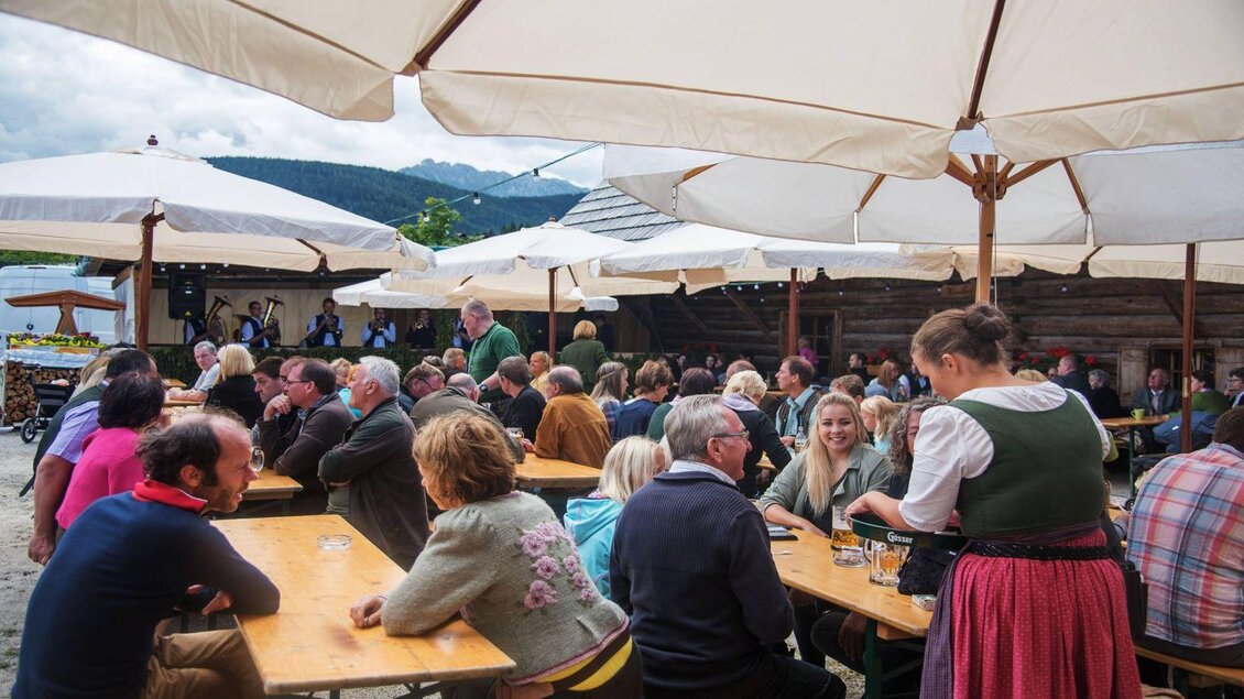 Eine große Gruppe von Menschen sitzt an langen Holztischen unter Sonnenschirmen. Die Atmosphäre ist festlich und es gibt traditionelle bayerische Kleidung. | © Anatevka Photography