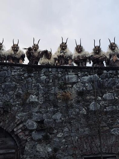 Group of masked figures with horns on a stone wall. They are wearing fur-like costumes and looking at the camera. | © Zeiritz Teifln