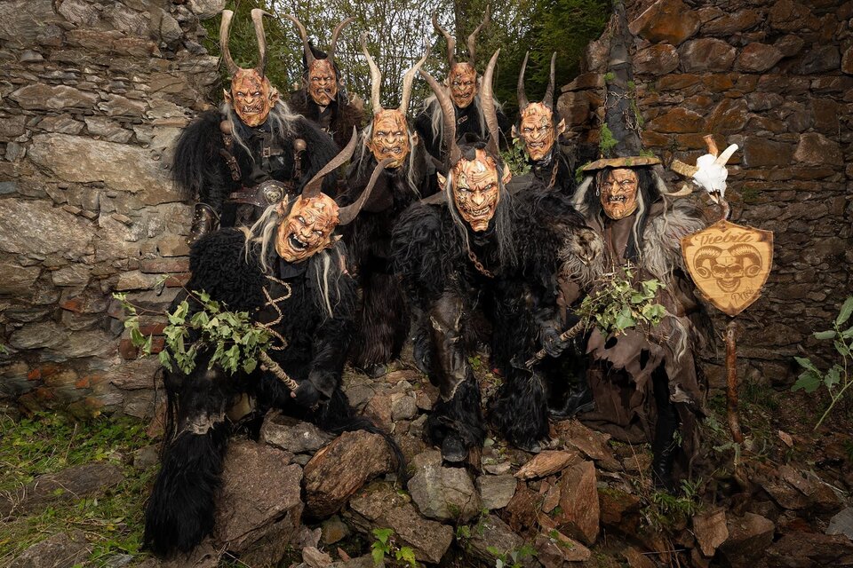 Gruppe von Maskenträgern in traditionellen kostümen, die auf einem felsigen Untergrund stehen. Sie tragen furchterregende Masken und sind von natürlichen Elementen umgeben. | © Trebiti Pass