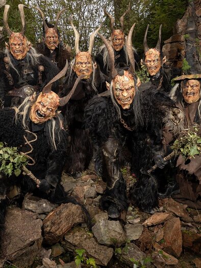 Gruppe von Maskenträgern in traditionellen kostümen, die auf einem felsigen Untergrund stehen. Sie tragen furchterregende Masken und sind von natürlichen Elementen umgeben. | © Trebiti Pass