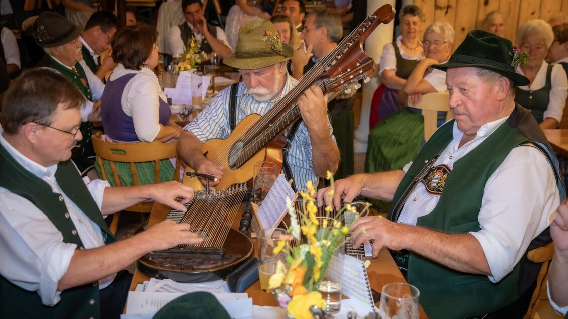 Eine gesellige Runde in einem traditionellen Wirtshaus. Drei Musiker spielen auf verschiedenen Instrumenten, während die Gäste im Hintergrund feiern. | © Manfred Digruber