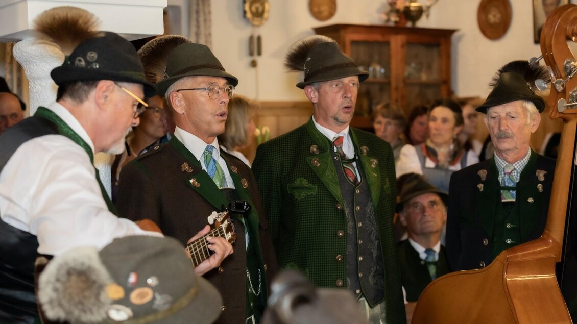 Eine Gruppe von Männern in traditionellen Trachten singt und musiziert in einer gemütlichen Atmosphäre. Im Hintergrund sind weitere Personen und eine rustikale Einrichtung zu sehen. | © Manfred Digruber