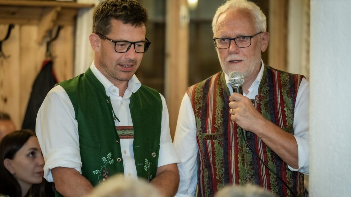 Zwei Männer in traditionellen Trachten stehen nebeneinander und sprechen in ein Mikrofon. Im Hintergrund sind weitere Personen zu sehen, die aufmerksam zuhören. | © Manfred Digruber