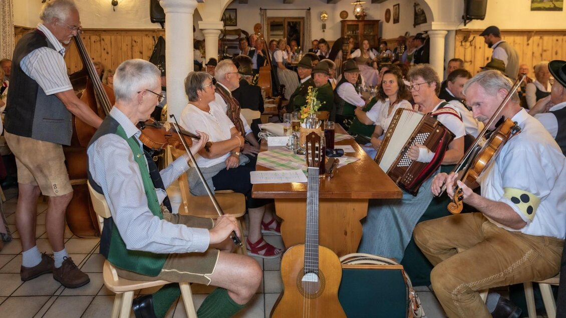 Eine gemütliche Gaststätte mit vielen Gästen, die traditionelle bayerische Musik genießen. Instrumente wie Geige, Kontrabass und Akkordeon sind zu sehen. | © Manfred Digruber