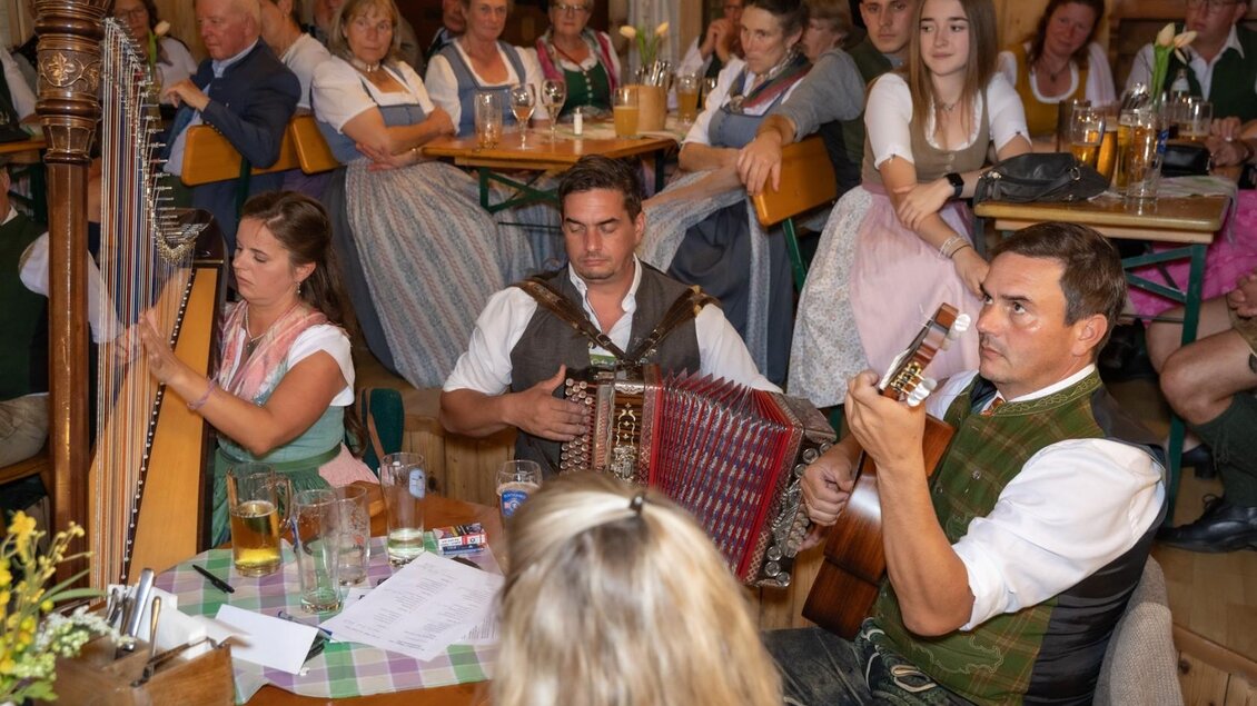 Eine traditionelle Veranstaltung mit Musikern, die Harfe, Akkordeon und Gitarre spielen. Im Hintergrund sitzen Gäste in Dirndln und Lederhosen, die die Darbietung genießen. | © Manfred Digruber