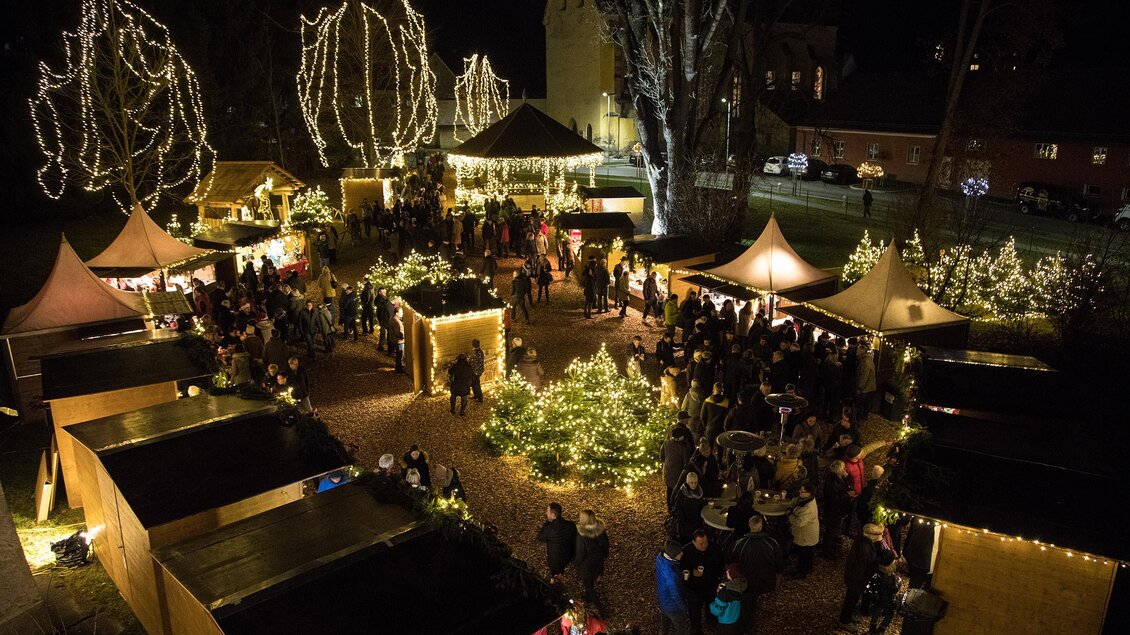Ein festlicher Weihnachtsmarkt bei Nacht mit beleuchteten Ständen und Bäumen. Viele Menschen genießen die festliche Atmosphäre und die Lichter. | © Harald Tauderer Photography