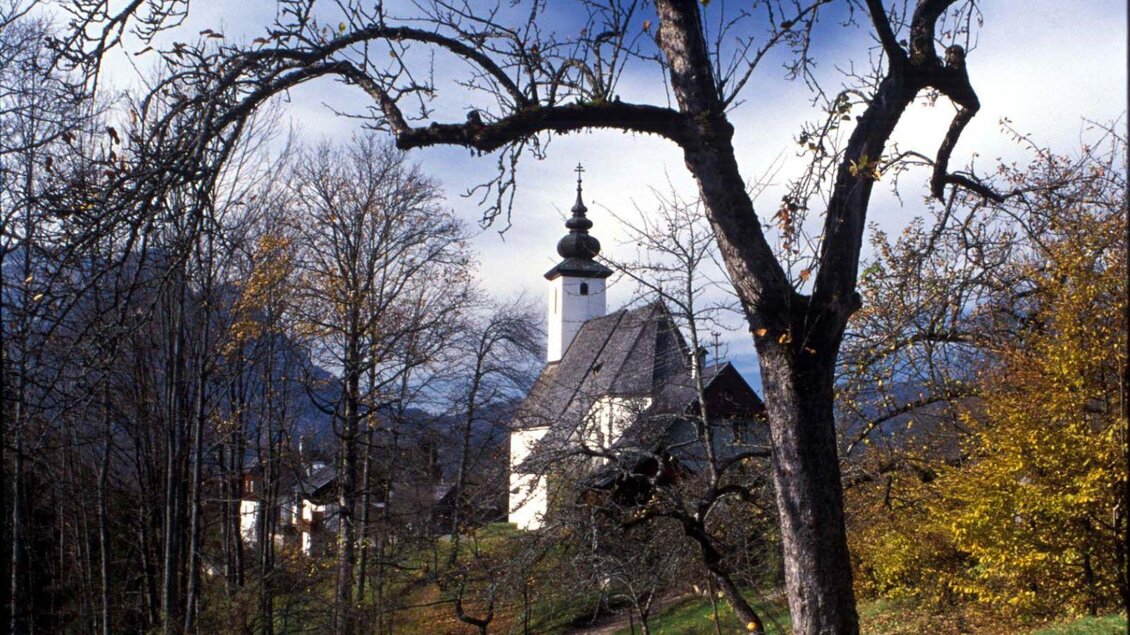 Ein roter Weihnachtsbaumkugel hängt an einem schneebedeckten Baum. Im Hintergrund leuchten festliche Lichter eines Weihnachtsmarktes. | © Tourismusverband Bad Aussee