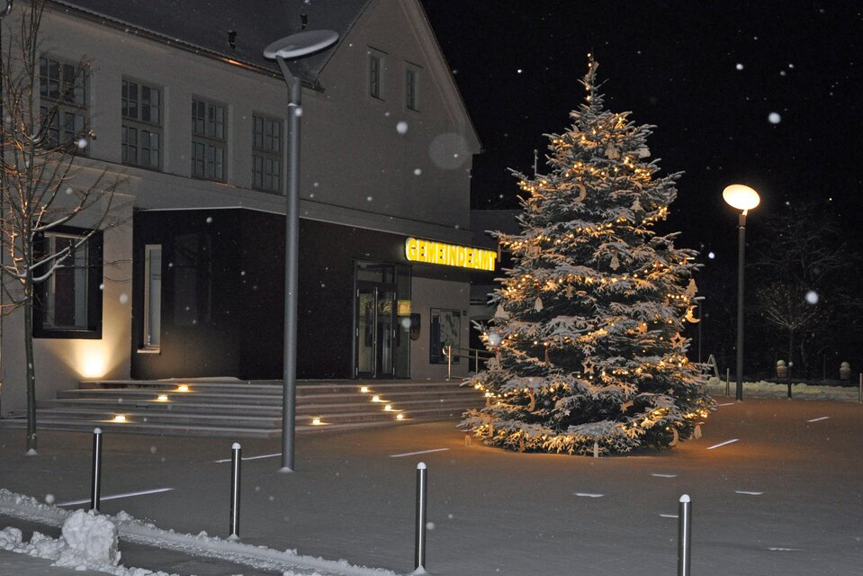 Ein festlich beleuchteter Weihnachtsbaum steht vor einem Gebäude. Es schneit leicht und die Umgebung ist in winterlicher Stimmung. | © Gemeinde Bad Loipersdorf