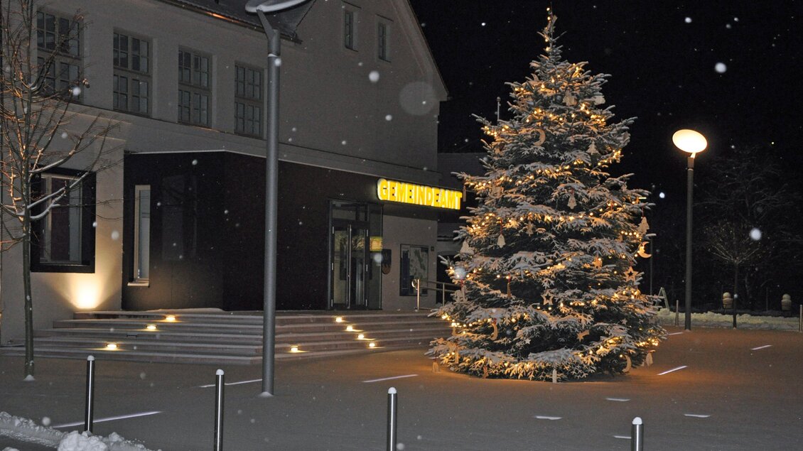 Ein festlich beleuchteter Weihnachtsbaum steht vor einem Gebäude. Es schneit leicht und die Umgebung ist in winterlicher Stimmung. | © Gemeinde Bad Loipersdorf