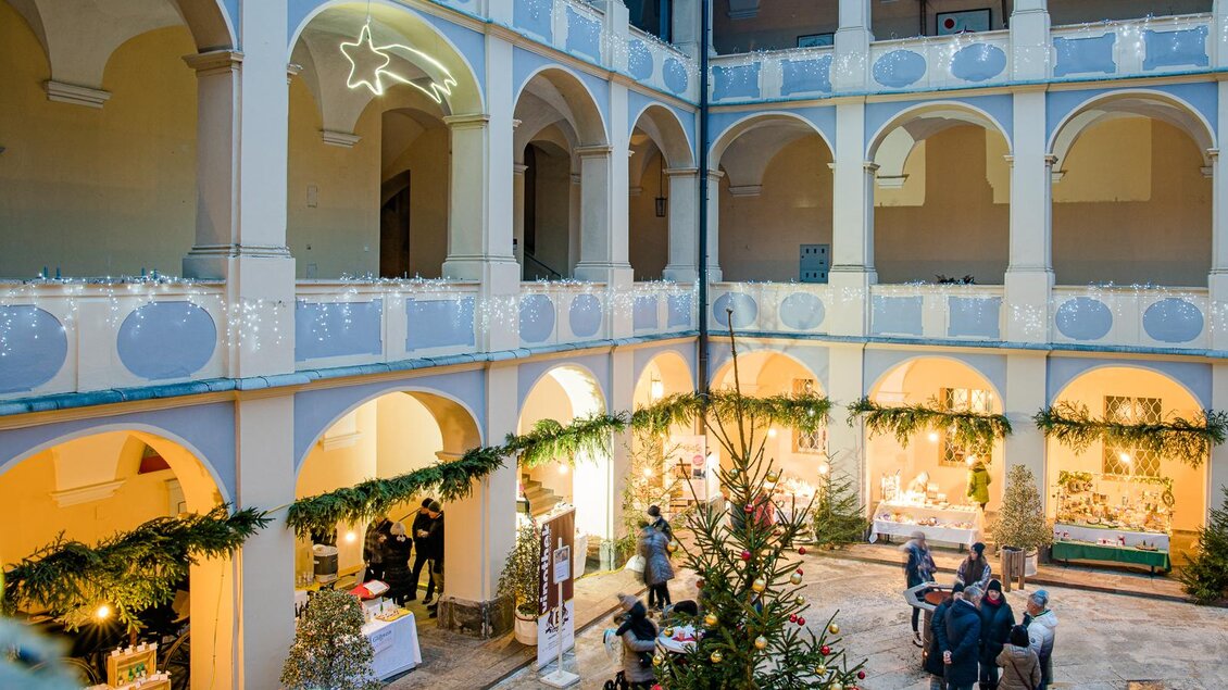 Adventmarkt im Arkadenhof von Schloss Piber mit geschmücktem Weihnachtsbaum, beleuchteten Marktständen und Besuchern. | © Region Graz-Harry Schiffer