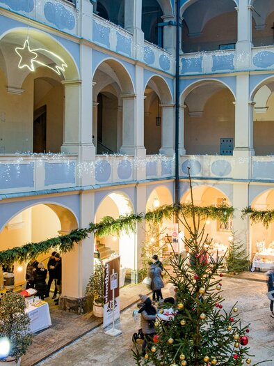 Adventmarkt im Arkadenhof von Schloss Piber mit geschmücktem Weihnachtsbaum, beleuchteten Marktständen und Besuchern. | © Region Graz-Harry Schiffer