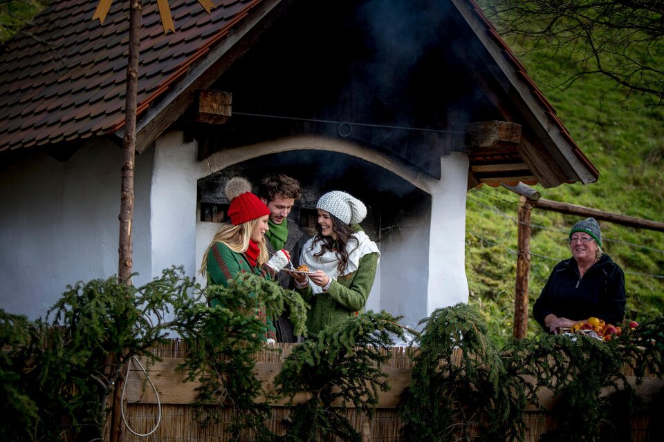Drei Besucher genießen eine Kostprobe vor einem Holzofen beim Adventmarkt am Hüblerhof in Semriach. | © Region Graz-Tom Lamm