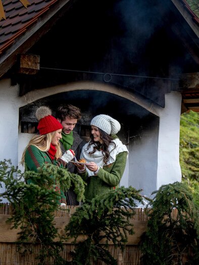 Drei Besucher genießen eine Kostprobe vor einem Holzofen beim Adventmarkt am Hüblerhof in Semriach. | © Region Graz-Tom Lamm