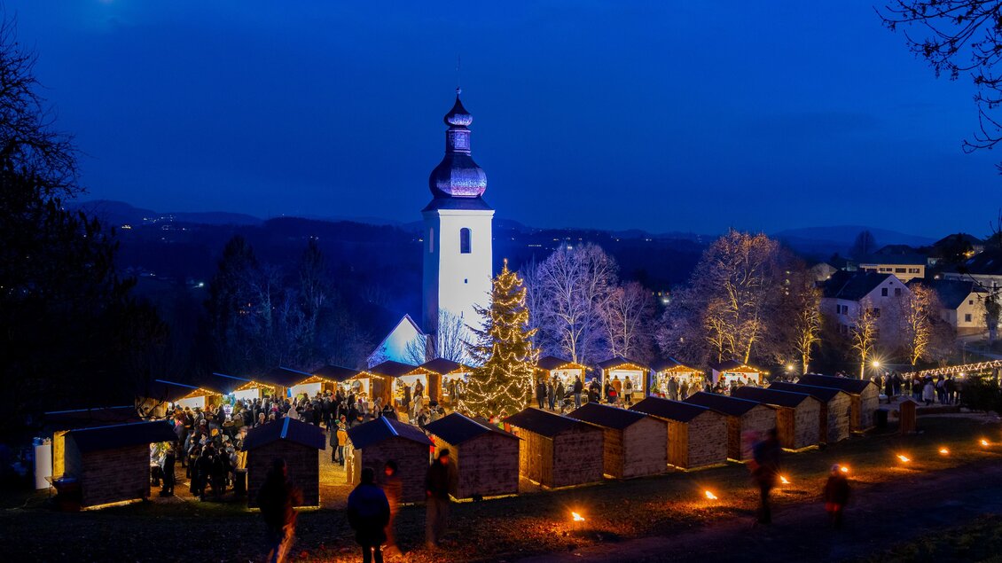 Beleuchtete Adventhütten, ein Weihnachtsbaum und zahlreiche Besucher vor der Kirche beim Barthlmäer Advent in der Dämmerung. | © Region Graz-Harry Schiffer