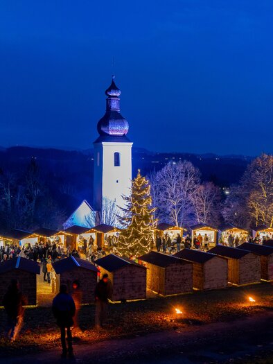 Illuminated Advent huts, a Christmas tree, and numerous visitors in front of the church at the Barthlmäer Advent during twilight. | © Region Graz-Harry Schiffer